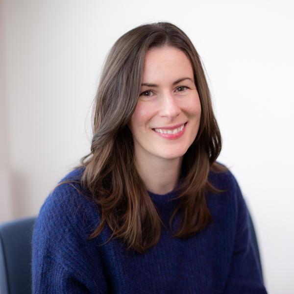 A smiling women with medium length dark hair in a blue sweater is seated on a dark blue chair.