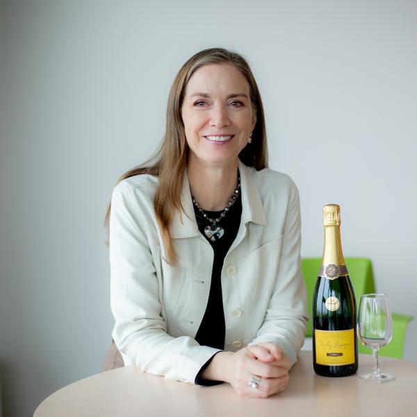 A women with shoulder length medium auburn hair sits smiling at a table smiling with a bottle and champagne glass next to her. 
