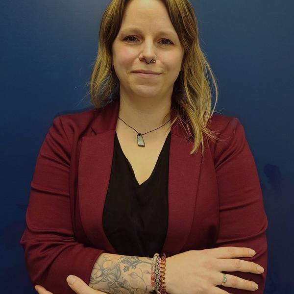 A close up of a woman smiling with her arms crossed with shoulder length blond hair in a burgundy blazer and black v-neck.