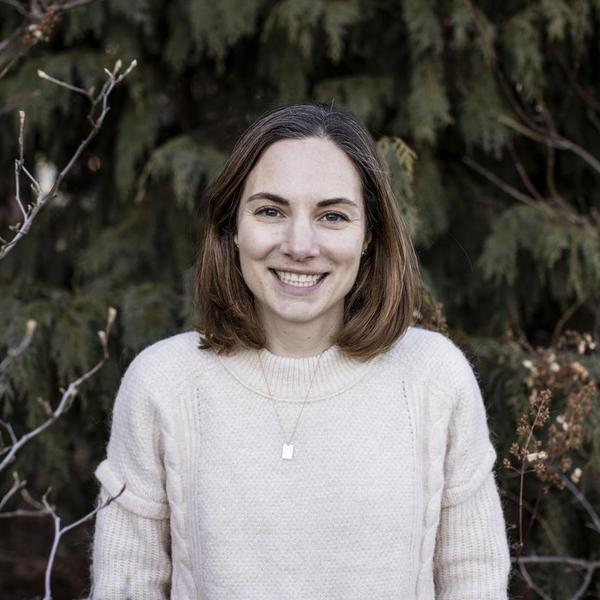 woman smiling in front of trees in a pink sweater and shoulder length brown hair. 