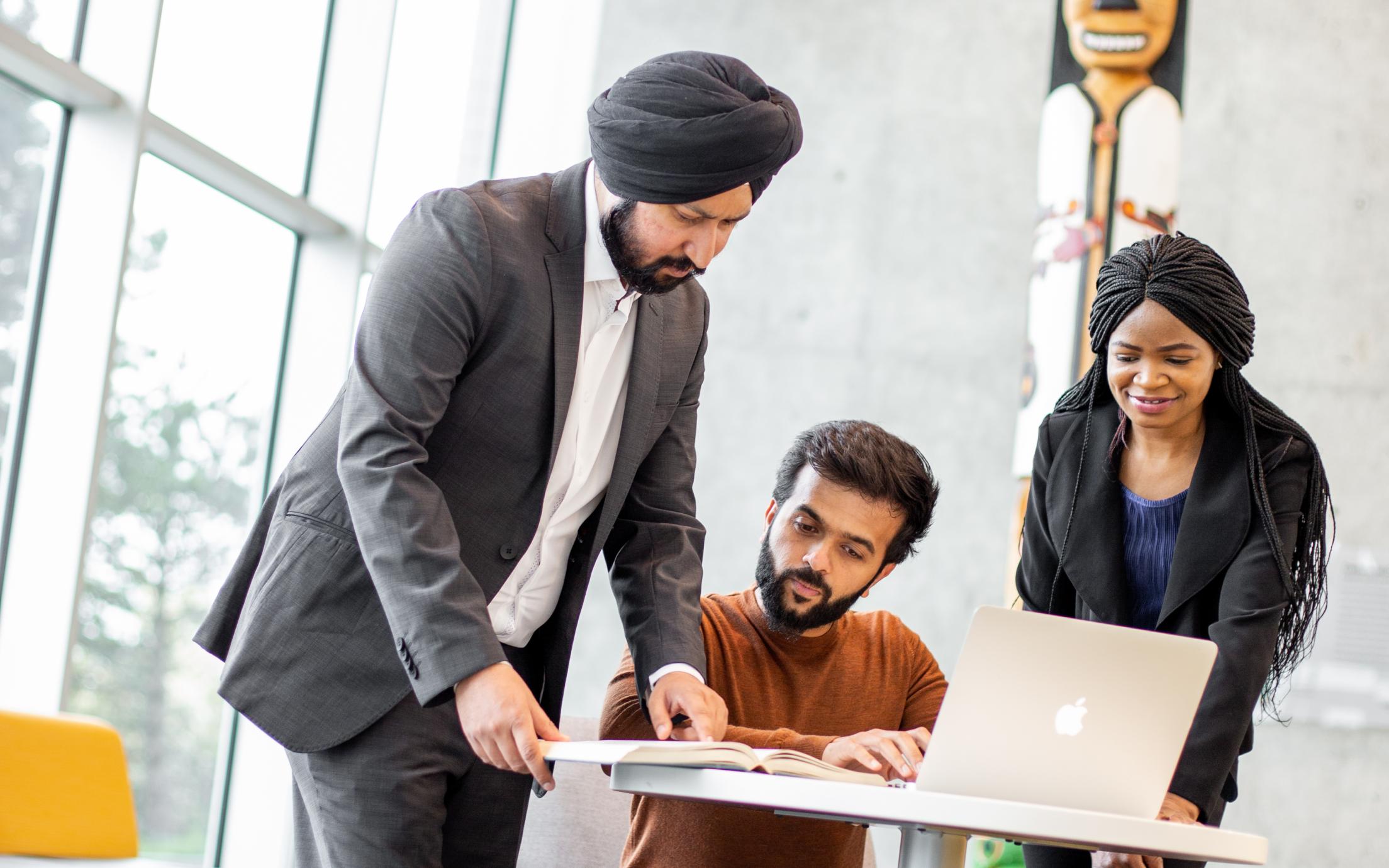 A group of 3 diverse, mature students are at a table with a laptop, working in s room with large, bright windows an a traditional totem in the background