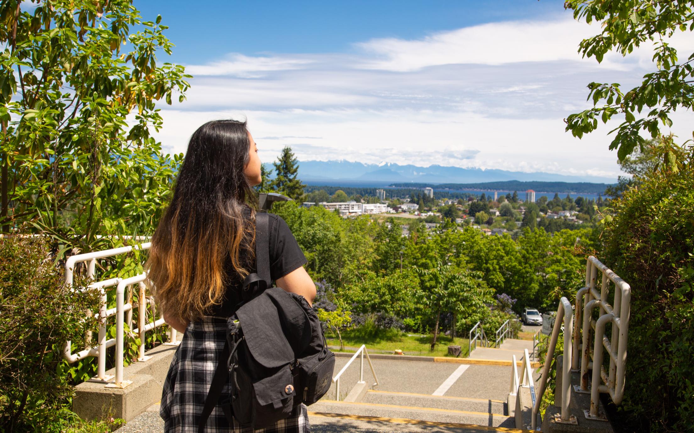 A young-adult standing at the top of stairs with long dark hair and a black backpack and a plaid shirt tied around her waist looking out at the trees and city in the distance.