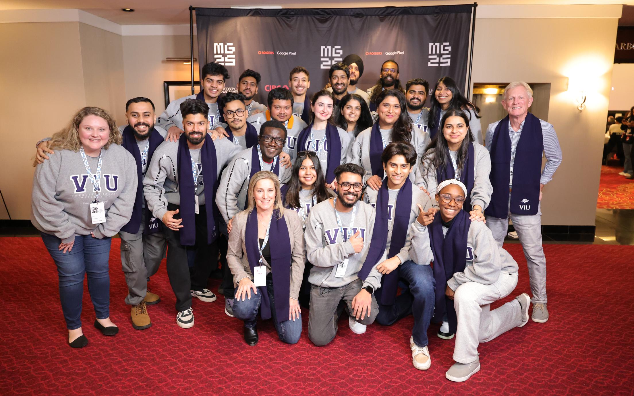 A group of 20 multicultural and multinational MBA students with three faculty coaches crouching in three rows with a the MBA games 25 sign background