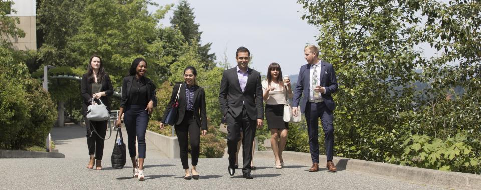 Six students dressed in professional attire walking on a cement path with trees in the background.