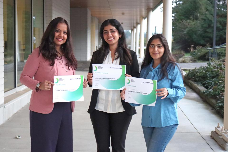Three smiling female students with long dark hair in casual professional attire holding their DMI certificates