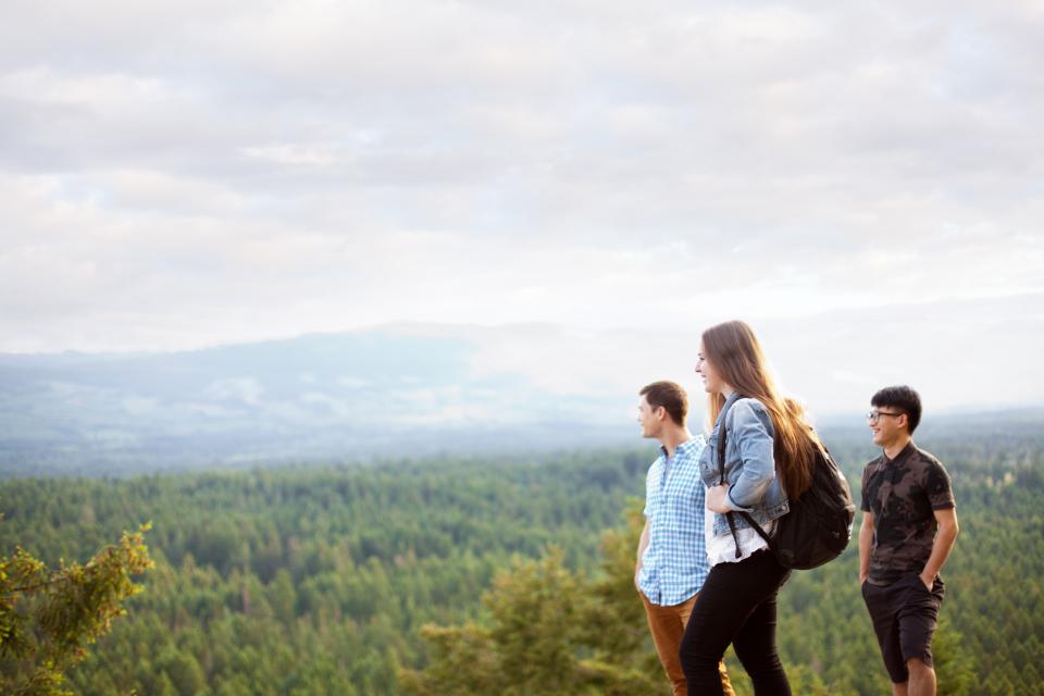 Three students, one with a backpack, dressing in casual clothing standing on top of a mountain looking out a forest.