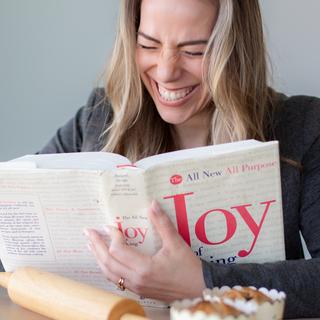 A blond woman laughing with her eyes closed holding a cook book.