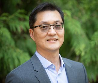 A man with short dark hair in glasses, a grey suit jacket and blue dress shirt smiling in from of some green trees.
