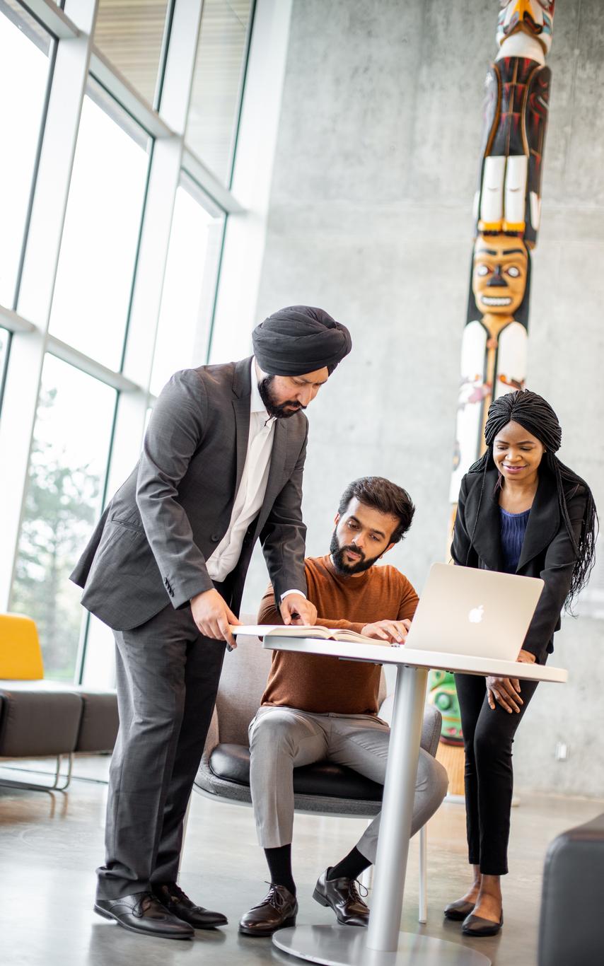 A group of 3 diverse, mature students are at a table with a laptop, working in s room with large, bright windows an a traditional totem in the background