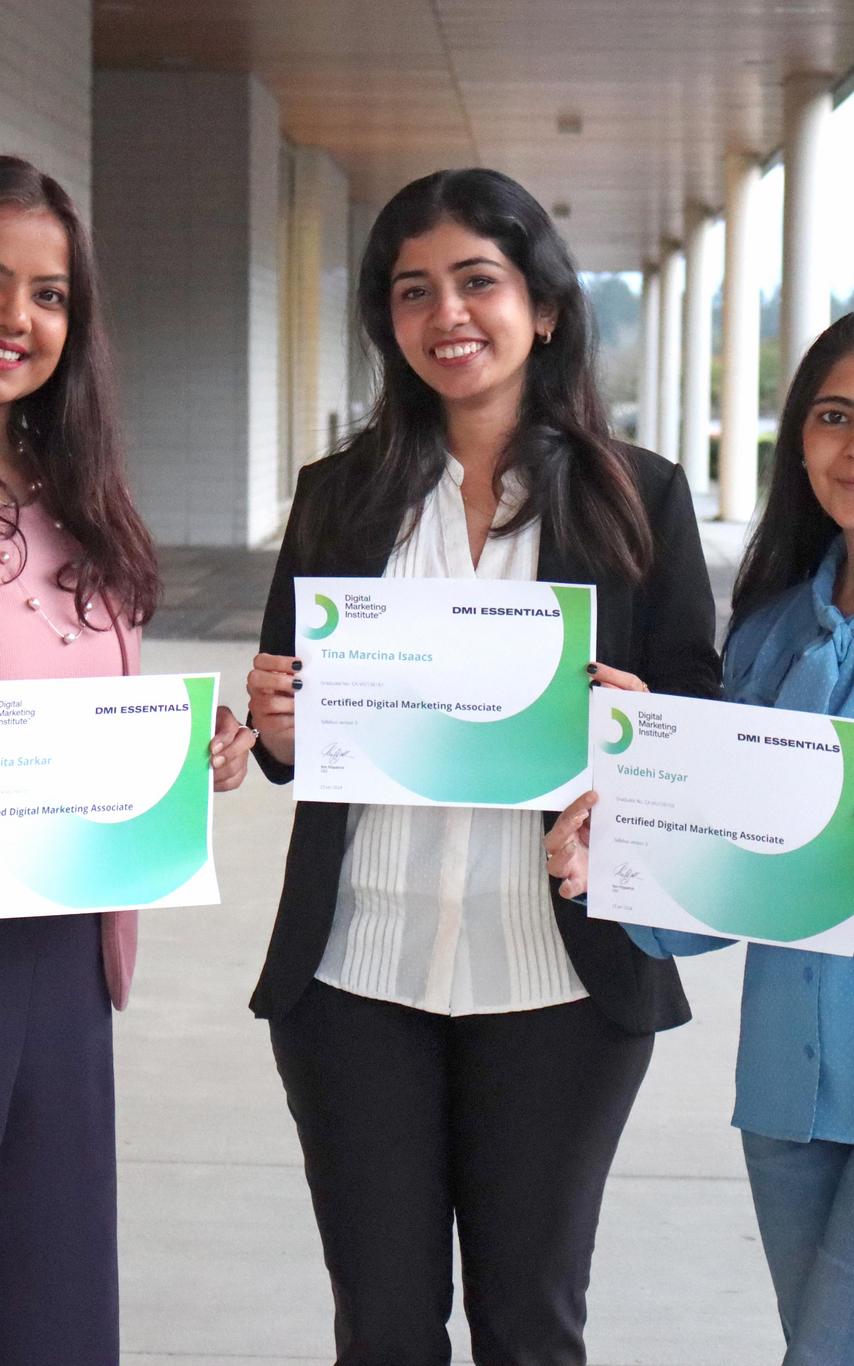 Three smiling female students with long dark hair in casual professional attire holding their DMI certificates