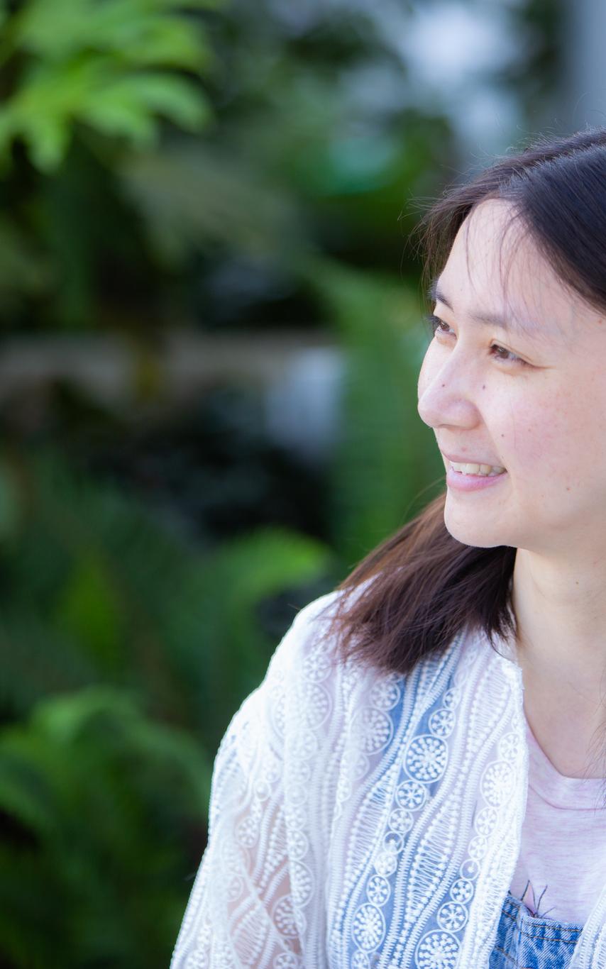 Female student with with shoulder length dark hair smiling and looking left in front of some trees.