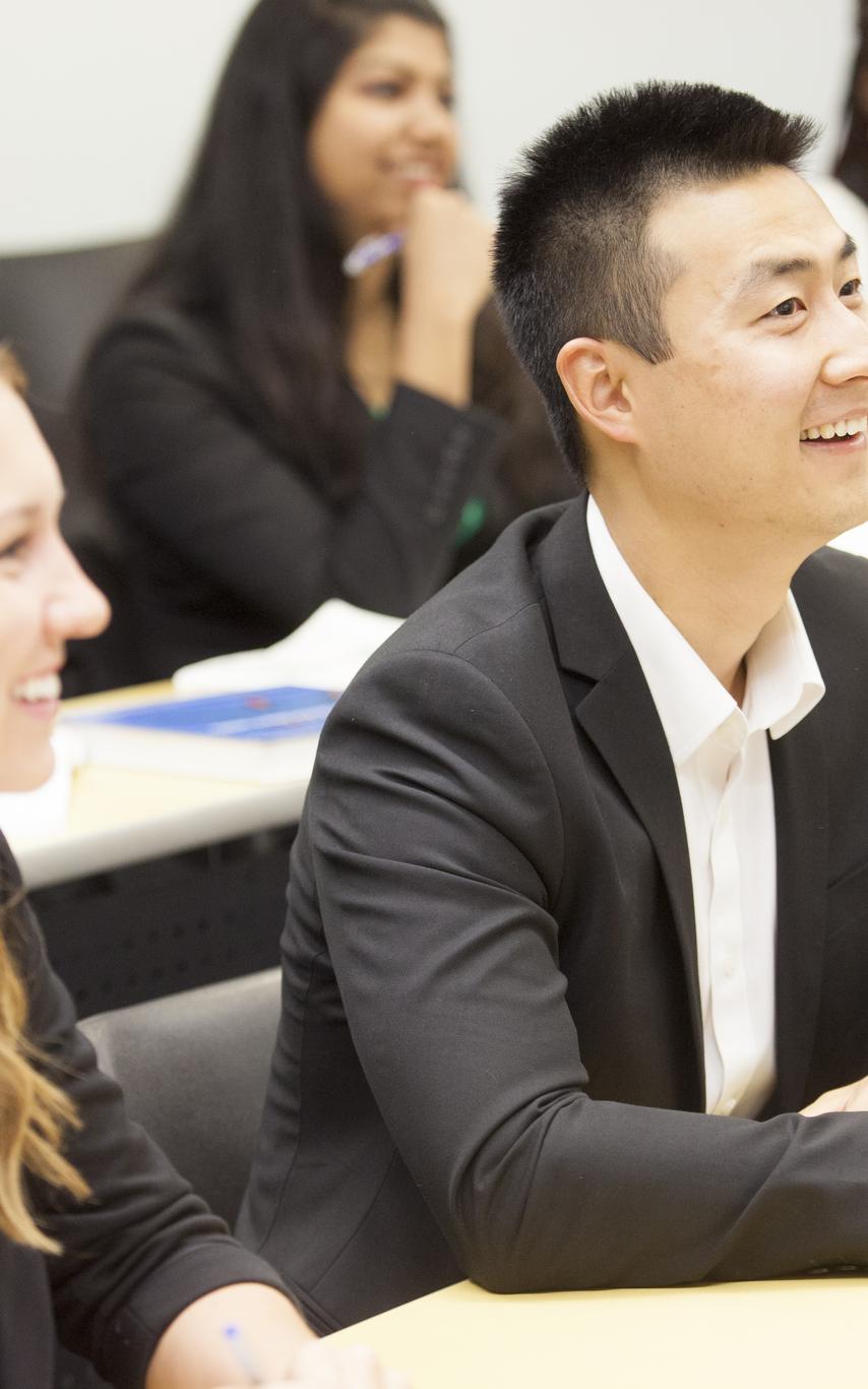 Five smiling students from divers backgrounds, dressed professionally, sitting at long rows in a classroom.
