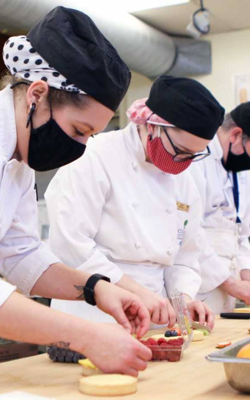 Three culinary students in masks, white culinary uniforms and black hats at a tall work table arranging food.