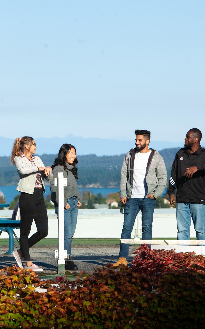 four students leaning on a railing on a rooftop patio with climbing leaves in the foreground and a person at a table and a mountain in the background