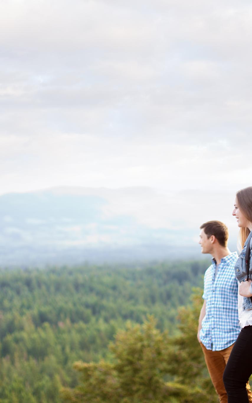 Three students, one with a backpack, dressing in casual clothing standing on top of a mountain looking out a forest.